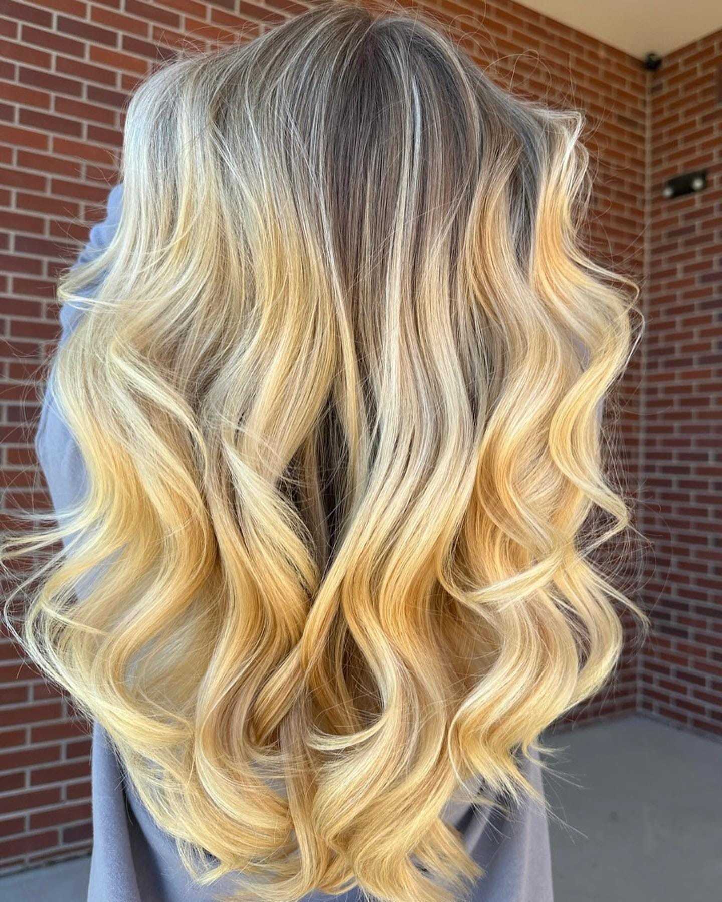 Woman with long, wavy balayage hair standing against brick wall.